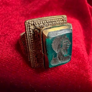 May include: An antique gold ring with a rectangular, green stone. The stone has a carved profile of a person. The ring has detailed patterns on the band and setting, displaying a vintage style.