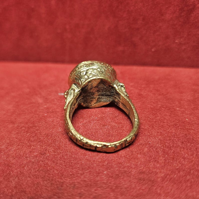 May include: A gold-colored ring with an oval-shaped stone. The ring has intricate detailing around the stone and along the band. The ring is set against a red background.