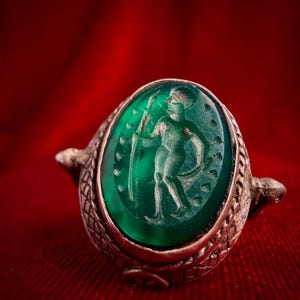 Op de afbeelding: Een zilveren ring met een ovale, doorschijnende groene edelsteen waarin een figuur met een staf is gegraveerd. De metalen band heeft een textuur met een ruitpatroon dat op schubben lijkt, met kleine dierachtige koppen aan de zijkanten.