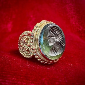 May include: A gold ring featuring an oval-shaped, translucent green stone engraved with a profile of a figure wearing a headdress. The metal band displays intricate, raised scrollwork and decorative patterns surrounding the central setting.