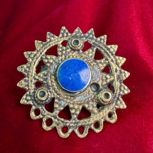 Puede incluir: Un broche circular de latón con un marco exterior texturizado en forma de sol, bordes dentados y detalles calados. Una piedra redonda, lisa y de color azul intenso está engastada en el centro, rodeada por tres pequeños acentos circulares oscuros.