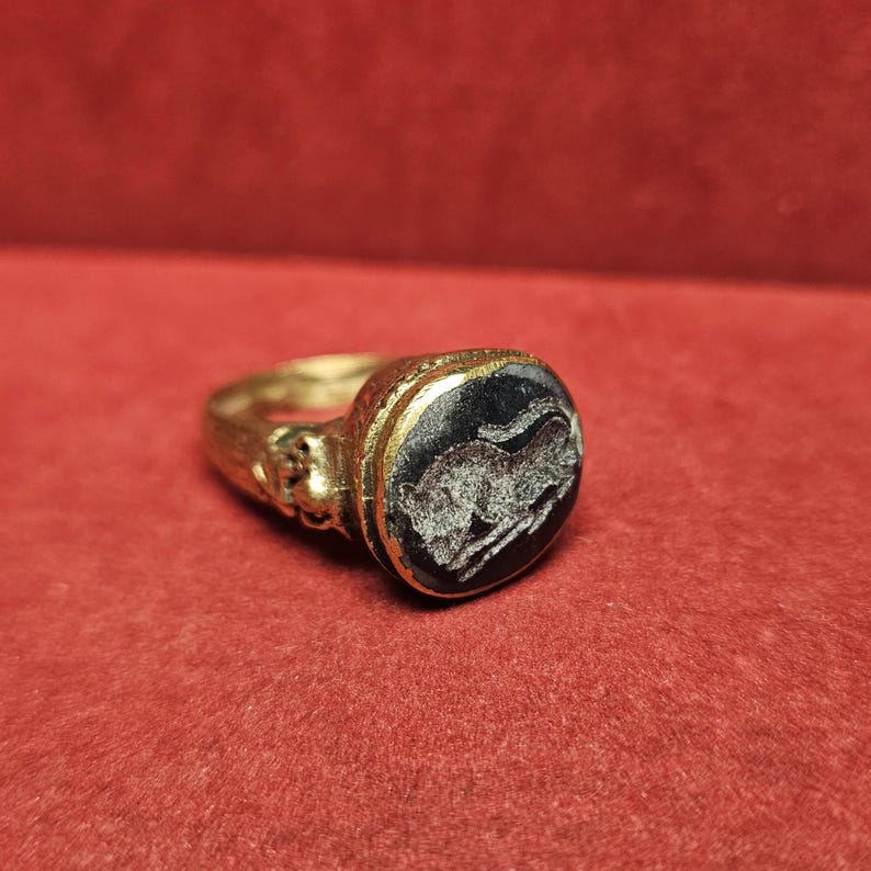 May include: A gold ring with an oval, black stone. The stone features a white, engraved image of an animal, possibly a horse or bull. The ring has intricate detailing on the band.