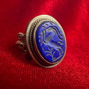 Peut inclure: Une bague en argent avec une pierre ovale en lapis-lazuli bleu profond gravée d'un cheval cabré. La monture métallique présente une bordure en corde torsadée et des volutes décoratives sur les côtés de l'anneau, formant un travail détaillé.