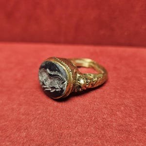 May include: A gold ring with a dark, possibly onyx, oval stone. The stone features a white, carved design. The ring's band has a textured, possibly antique, appearance. The ring is set against a red background.