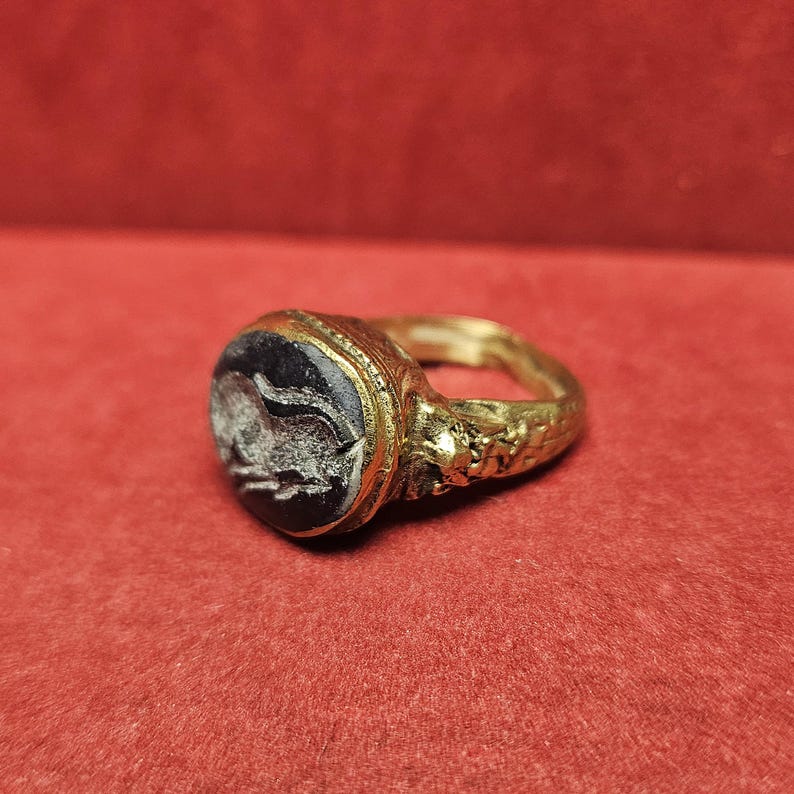 May include: A gold ring with a dark, possibly onyx, oval stone. The stone features a white, carved design. The ring's band has a textured, possibly antique, appearance. The ring is set against a red background.