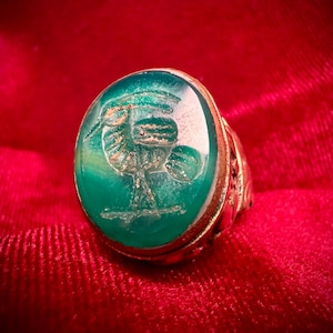 May include: A gold ring featuring a large, oval, green gemstone. The stone is intricately engraved with a design. The ring is set against a red velvet backdrop, providing a strong contrast. The ring's band has detailed patterns.