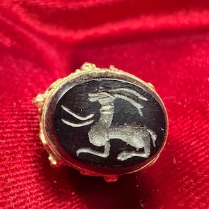 Puede incluir: Un anillo antiguo dorado con una piedra negra ovalada. La piedra presenta una imagen tallada de un ciervo o gacela. El anillo está sobre un fondo rojo, resaltando los detalles y el diseño del anillo. Una pieza de joyería única.