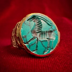 Puede incluir: Un anillo de oro con una incrustación de piedra redonda de color turquesa con una figura animal estilizada tallada. La piedra muestra vetas oscuras similares a una red en su superficie, y la banda de metal tiene un acabado texturizado y adornado.