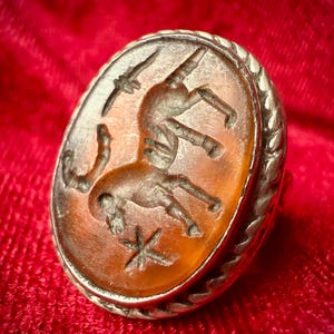 May include: Oval antique ring with a silver band and a carved amber stone. The stone features a detailed engraving of a figure riding an animal, a bird, and a star. The ring has a textured border.