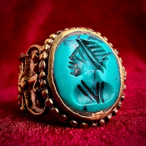 May include: A gold-toned ring featuring an oval, bright turquoise-coloured stone with an intaglio carving of a profile. The setting is decorated with a beaded border and ornate, textured metalwork along the sides of the band, creating a detailed look.