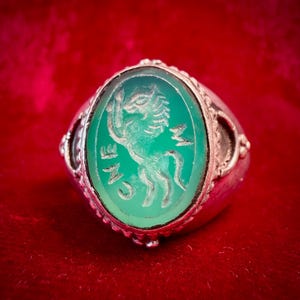 Könnte beinhalten: Ein Silberring mit einem ovalen, durchscheinenden grünen Stein in einer dekorativen Metallfassung. Der Stein ist mit einem steigenden Pferd und den Buchstaben CNE graviert, wobei die Seiten des Rings aufwendige Verzierungen aufweisen.