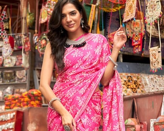 May include: A woman wearing a vibrant pink sari with a white floral pattern. The sari is draped elegantly, showcasing the intricate design. The background features a market scene with various bags and textiles.