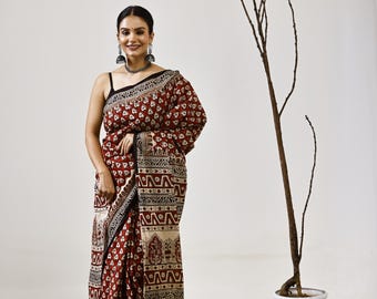 May include: A woman wearing a maroon and cream-colored sari with a geometric and floral pattern. The sari has a black border and is draped over the shoulders. The woman is wearing a black top and silver jewelry.