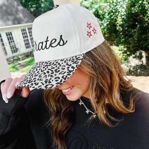 May include: White baseball cap with "Pirates" script and red star embroidery. The brim features a black and white leopard print. A stylish accessory for sports fans.