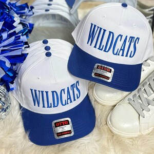 May include: Two white baseball caps with blue brims. Both caps have the word "WILDCATS" embroidered in blue on the front. The caps are on a white fuzzy surface.