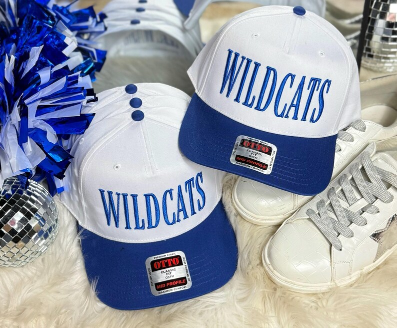 May include: Two white baseball caps with blue brims. Both caps have the word "WILDCATS" embroidered in blue on the front. The caps are on a white fuzzy surface.