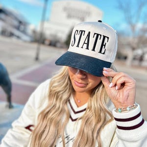 May include: A white and black baseball cap with the word "STATE" embroidered in black on the front. The cap is being worn by a person with long blonde hair.