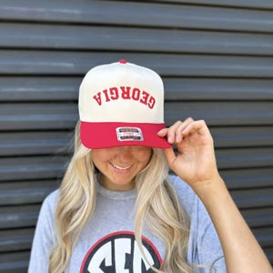 May include: A cream-colored baseball cap with a red brim and the word "GEORGIA" embroidered in red. The cap has a flat bill and a snapback closure. The cap is being held up by a person.