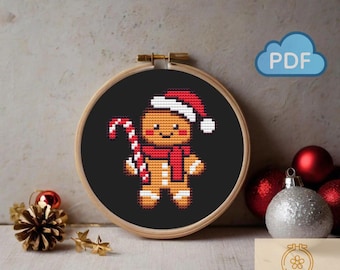 May include: A cross-stitch pattern of a gingerbread man wearing a Santa hat and scarf, holding a candy cane, set in a wooden embroidery hoop. The background is black. A PDF icon is in the upper right.