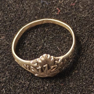 May include: A silver ring with the number "172" in the center, surrounded by a floral design. The ring is on a dark, textured surface.