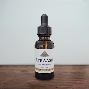 May include: A brown glass bottle of eye wash with a black dropper. The label reads "EYEWASH" and "100% RAW & PURE" with a volume of 1 fl oz (30ml). The brand name "ANCIENT RESONANCE" is also printed on the label. The bottle is on a wooden surface.