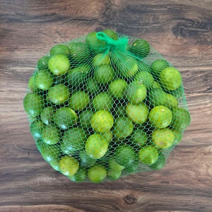 Puede incluir: Una bolsa de red verde llena de limas frescas. Las limas son de varios tonos de verde y amarillo, algunas muestran signos de maduración. La bolsa está atada con una cinta verde, sobre un fondo de madera.