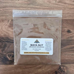 May include: A transparent plastic bag containing brown powder, labelled "MAYA NUT (Brosimum alicastrum)". The label states "100% RAW & PURE" and the weight is 113g. Instructions and a disclaimer are also printed on the label. The bag is on a wooden surface.