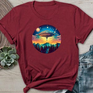 May include: Maroon t-shirt featuring a circular graphic of a UFO hovering over a mountain landscape with a starry sky. The design includes a UFO, trees, mountains, and a colorful sunset. The shirt is displayed with a watch, succulent, and jeans.