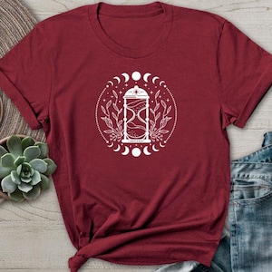 May include: A maroon t-shirt with a white graphic of an hourglass with moon phases and floral accents.