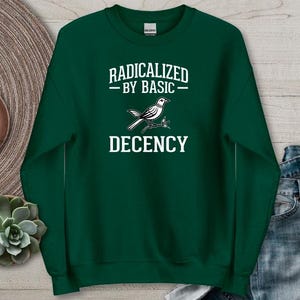 May include: A forest green sweatshirt featuring the words "RADICALIZED BY BASIC DECENCY" and a bird graphic. The design is in white. The sweatshirt is made of a soft material and has a crew neck.