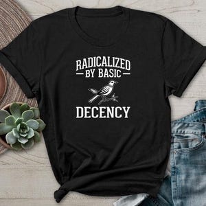Puede incluir: Camiseta negra con el texto blanco "RADICALIZED BY BASIC DECENCY" y una ilustración de un pájaro. La camiseta se muestra sobre una superficie de madera con elementos decorativos.