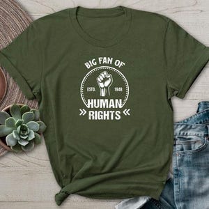 Puede incluir: Camiseta verde militar con un gráfico blanco que dice "BIG FAN OF HUMAN RIGHTS" alrededor de un puño en alto. El texto incluye "ESTD. 1948". La camiseta se muestra sobre una superficie de madera con un reloj y una planta suculenta.