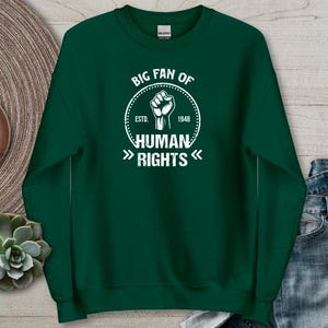 Puede incluir: Sudadera verde bosque con el texto blanco "BIG FAN OF HUMAN RIGHTS". El diseño incluye un puño en alto dentro de un círculo, con "ESTD. 1948" alrededor. La sudadera se muestra sobre una superficie neutra con un reloj y una suculenta.