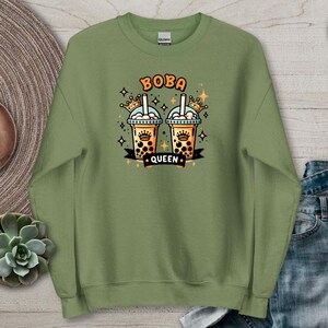 Może przedstawiać: Zielona wojskowa bluza z nadrukiem dwóch napojów boba tea, każdy z koroną, oraz napisem "BOBA QUEEN". Projekt zawiera gwiazdy i czarny baner. Bluza jest prezentowana na drewnianej powierzchni z elementami dekoracyjnymi.