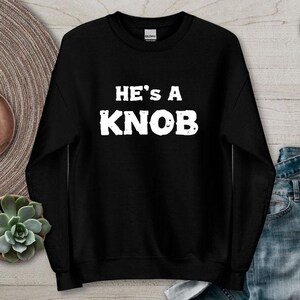 May include: Black sweatshirt with the text "HE'S A KNOB" in white, distressed lettering. The sweatshirt is displayed on a light-coloured surface with decorative items, including a succulent plant and earrings.