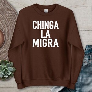 Könnte beinhalten: Dunkel schokoladenbraunes Sweatshirt mit dem weißen Text "CHINGA LA MIGRA". Das Sweatshirt wird auf einer Holzoberfläche mit einem dekorativen Teller, Ohrringen und einer Sukkulentenpflanze präsentiert.