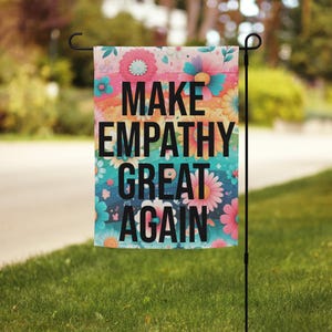 May include: A garden flag with the text "MAKE EMPATHY GREAT AGAIN" in black block letters. The flag has a floral design with pink, orange, blue, and white flowers. The flag is hanging on a black metal stand in a grassy yard.
