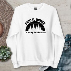 Może przedstawiać: Biała bluza z napisem "DIGITAL NOMAD" nad sylwetką lasu i frazą "I'm on My Own Deadline" w kolorze czarnym. Bluza jest prezentowana na jasnej powierzchni z zegarkiem i rośliną sukulent.