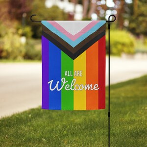 May include: A garden flag with a rainbow design and the words "ALL ARE Welcome" in white script. The flag has a black metal pole and is displayed on a green lawn.