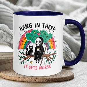 Pu&ograve; includere: Tazza da caff&egrave; bianca con bordo e manico blu. La tazza presenta un'immagine di un cartone animato di uno scheletro che tiene una tazza di caff&egrave; e un gatto nero seduto su un ramo. Il testo sulla tazza dice "Hang in there it gets worse".