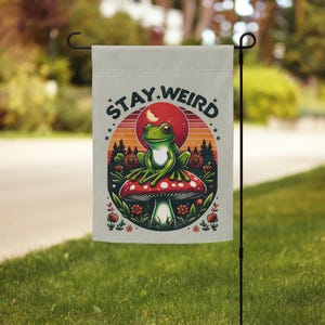 Puede incluir: Una bandera de jardín con fondo blanco y el texto "Stay Weird" en letras negras. Una rana verde se sienta en un hongo rojo y blanco con una puesta de sol y una luna en el fondo.