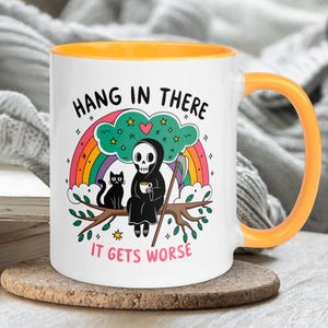 Pu&ograve; includere: Una tazza da caff&egrave; in ceramica bianca con un bordo e un manico gialli. La tazza presenta un'immagine di un cartone animato di uno scheletro che tiene una tazza di caff&egrave;, un gatto nero e un arcobaleno. Il testo sulla tazza dice "Hang in there it gets worse".