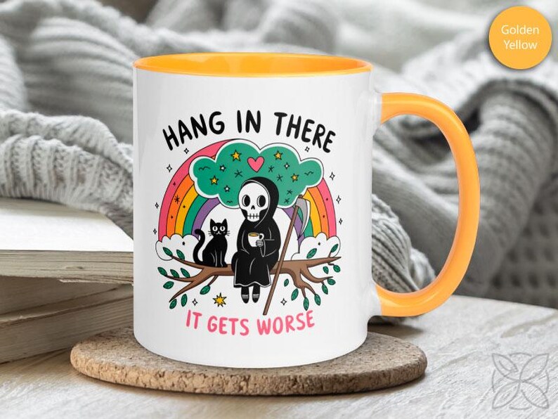 Pu&ograve; includere: Una tazza da caff&egrave; in ceramica bianca con un bordo e un manico gialli. La tazza presenta un'immagine di un cartone animato di uno scheletro che tiene una tazza di caff&egrave;, un gatto nero e un arcobaleno. Il testo sulla tazza dice "Hang in there it gets worse".