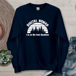 Może przedstawiać: Granatowa bluza z napisem "DIGITAL NOMAD" nad grafiką lasu i napisem "I'm on My Own Deadline". Bluza jest prezentowana na neutralnej powierzchni z zegarkiem i rośliną sukulent.