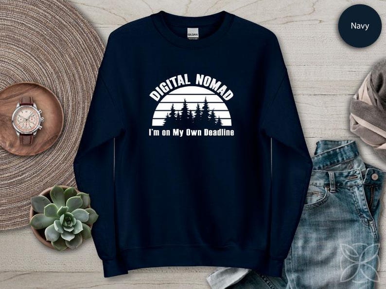 Może przedstawiać: Granatowa bluza z napisem "DIGITAL NOMAD" nad grafiką lasu i napisem "I'm on My Own Deadline". Bluza jest prezentowana na neutralnej powierzchni z zegarkiem i rośliną sukulent.