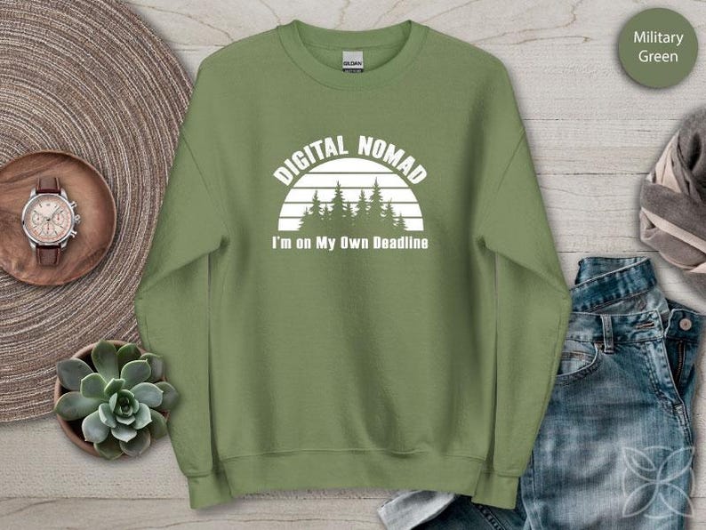 Może przedstawiać: Zielona wojskowa bluza z napisem "DIGITAL NOMAD" nad grafiką zachodu słońca i drzew. Poniżej grafiki znajduje się napis "I'm on My Own Deadline". Bluza jest prezentowana na drewnianej powierzchni.