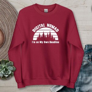 Może przedstawiać: Bordowa bluza z napisem "DIGITAL NOMAD" nad grafiką zachodu słońca i drzew, z napisem "I'm on My Own Deadline" poniżej. Bluza jest prezentowana na neutralnej powierzchni z zegarkiem i rośliną sukulent.