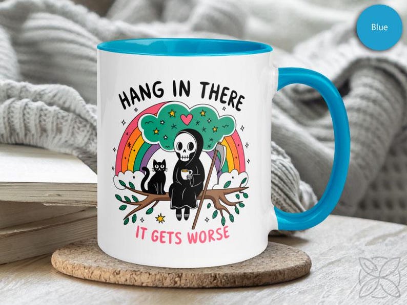 Pu&ograve; includere: Tazza da caff&egrave; in ceramica bianca con bordo e manico blu. La tazza presenta un'immagine di un cartone animato di uno scheletro che indossa una tunica con cappuccio, seduto su un ramo con un gatto. Il testo sulla tazza dice "Hang in there it gets worse".