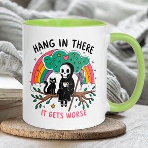 Pu&ograve; includere: Una tazza da caff&egrave; in ceramica bianca con un bordo e un manico verdi. La tazza presenta un'illustrazione di cartone animato di un Grim Reaper con un gatto nero seduto su un ramo con un arcobaleno dietro di loro. Il testo sulla tazza dice "Hang in there it gets worse".