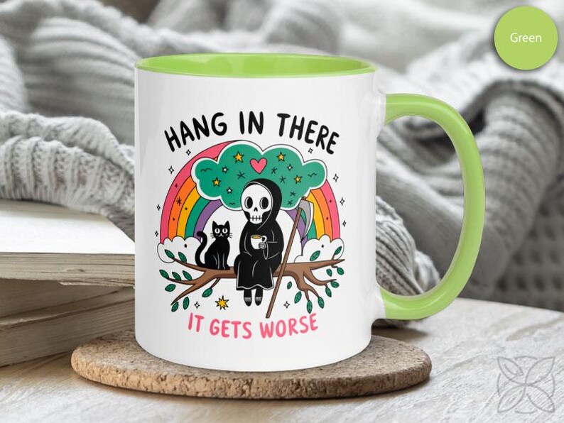 Pu&ograve; includere: Una tazza da caff&egrave; in ceramica bianca con un bordo e un manico verdi. La tazza presenta un'illustrazione di cartone animato di un Grim Reaper con un gatto nero seduto su un ramo con un arcobaleno dietro di loro. Il testo sulla tazza dice "Hang in there it gets worse".